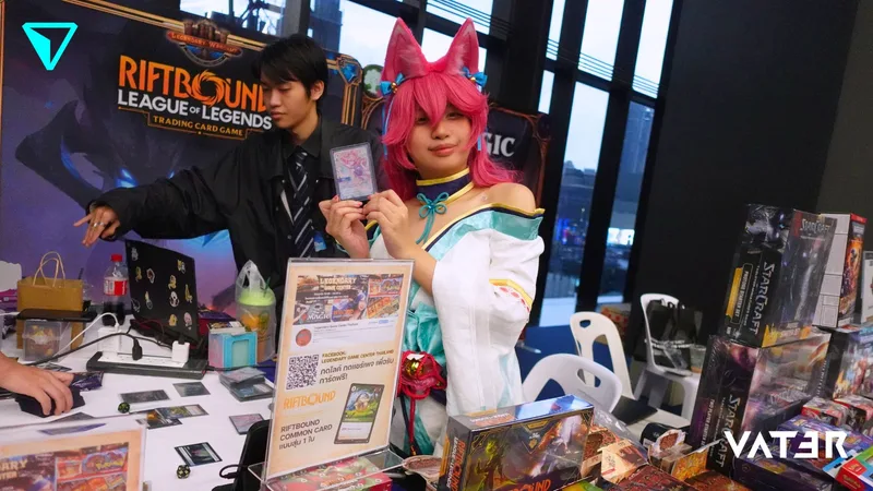 คอสเพลเยอร์สวมหูจิ้งจอกสีชมพูชูการ์ดเกมข้างพนักงานที่บูท Riftbound: League of Legends