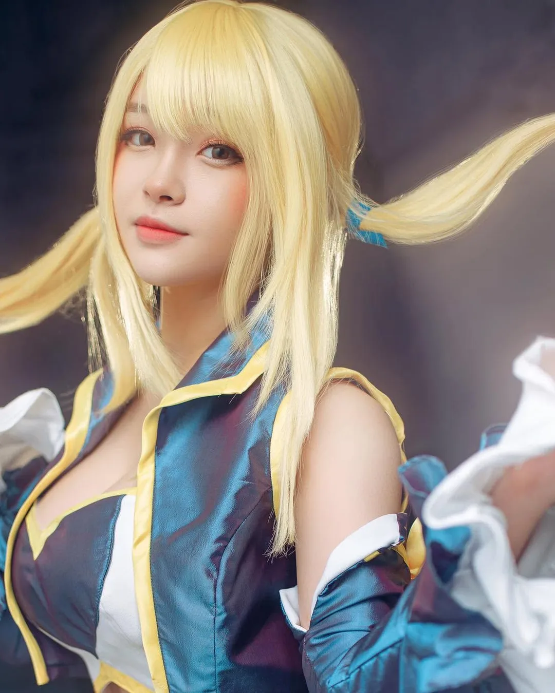 คอสเพลเยอร์แต่งกายเป็น Lucy Heartfilia จาก Fairy Tail สวมวิกผมทวินเทลสีบลอนด์และชุดสีน้ำเงินขาว กำลังจับผมโพสท่ามองกล้อง