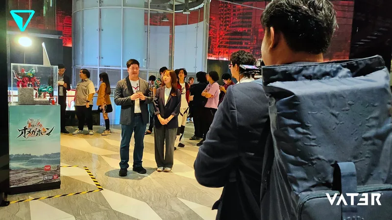 พิธีกรบรรยายต่อหน้าผู้ร่วมงานใกล้ตู้โชว์ฟิกเกอร์ในงานอีเวนต์โปรโมตเกมในร่ม