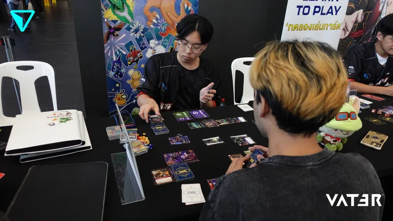 ชายหนุ่มสองคนนั่งเล่นการ์ดเกมด้วยกันที่โต๊ะภายในบูธงานอีเวนต์เกม