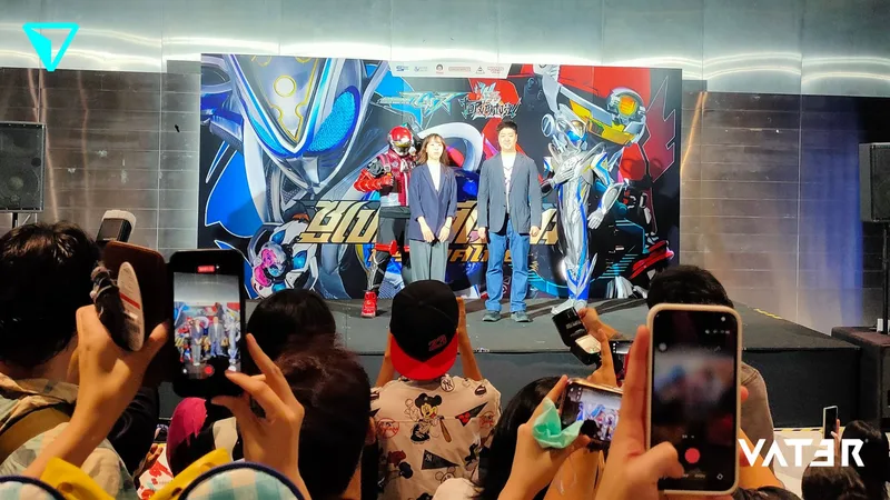 ผู้ชมใช้สมาร์ตโฟนถ่ายภาพขณะที่ตัวละครซูเปอร์ฮีโร่และผู้ร่วมงานสองคนยืนโพสท่าบนเวทีหน้าป้ายแบ็กดรอป