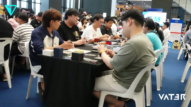 ผู้เล่นนั่งเล่นการ์ดเกมรอบโต๊ะระหว่างการแข่งขันภายในฮอลล์จัดงานที่มีผู้คนมากมาย