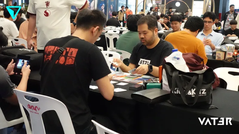 ผู้เข้าร่วมแข่งขันนั่งประจันหน้ากันเล่นการ์ดเกมบนโต๊ะสีดำในงานแข่งขันเกมที่เต็มไปด้วยผู้คน