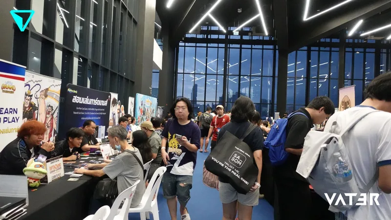 ผู้คนนั่งเล่นการ์ดเกมที่โต๊ะตามบูธ ขณะที่ผู้ร่วมงานคนอื่นๆ เดินชมในฮอลล์จัดงานที่มีกระจกบานใหญ่