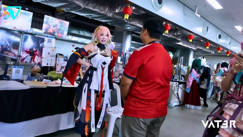 คอสเพลเยอร์ผมสีชมพูยืนอยู่ที่บูธในงานอีเวนต์และพูดคุยกับชายในเสื้อสีแดง