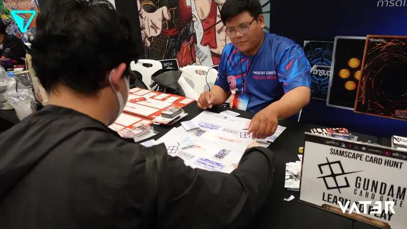 ชายสองคนนั่งตรงข้ามกันเล่น Gundam Card Game ที่โต๊ะบูธในงานกิจกรรม