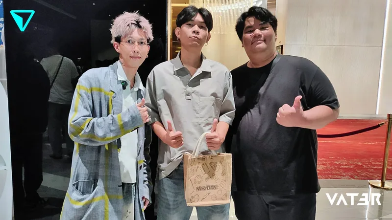 ชายหนุ่มสามคนยืนโพสท่าชูนิ้วโป้งร่วมกันในอาคาร โดยคนตรงกลางถือถุงของขวัญใบเล็ก