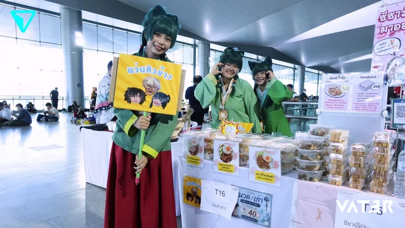 คอสเพลเยอร์เหมาเหมายิ้มอยู่หลังซุ้มขายอาหารในงานอนิเมะ โดยคนหนึ่งถือป้ายโปรโมต