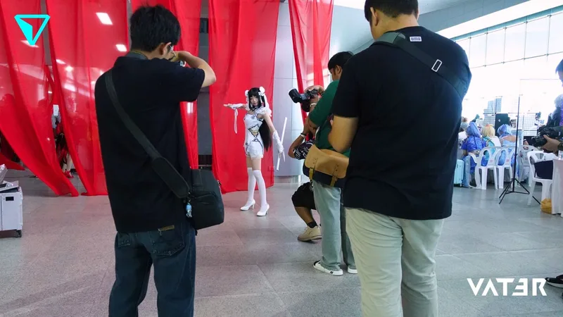 คอสเพลเยอร์ในชุดสีขาวกำลังโพสท่าหน้าผ้าม่านสีแดง ขณะที่ช่างภาพกำลังถ่ายรูปในงานจัดแสดง