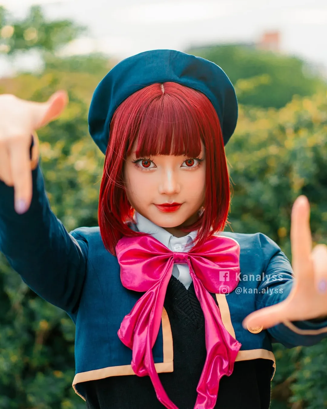 คอสเพลเยอร์ในชุด Kana Arima จาก Oshi no Ko ทำท่ามือเป็นกรอบภาพอยู่ด้านหน้าพุ่มไม้สีเขียว