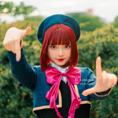 คอสเพลเยอร์ในชุด Kana Arima จาก Oshi no Ko ทำท่ามือเป็นกรอบภาพอยู่ด้านหน้าพุ่มไม้สีเขียว