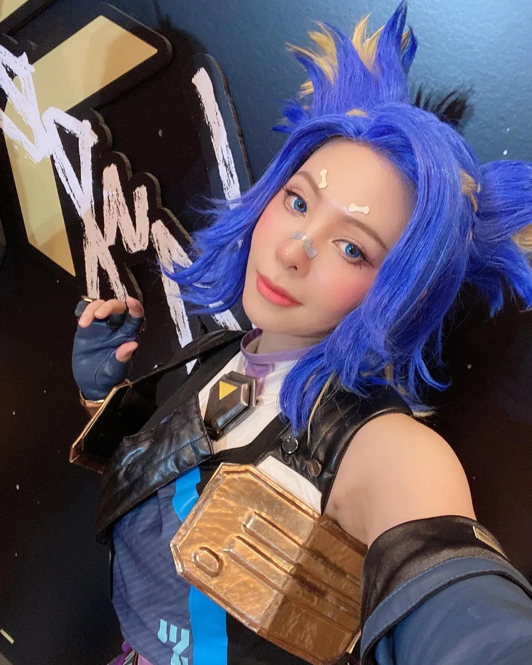 A cosplayer dressed as Neon from Valorant with bright blue hair and tactical attire takes a selfie indoors.