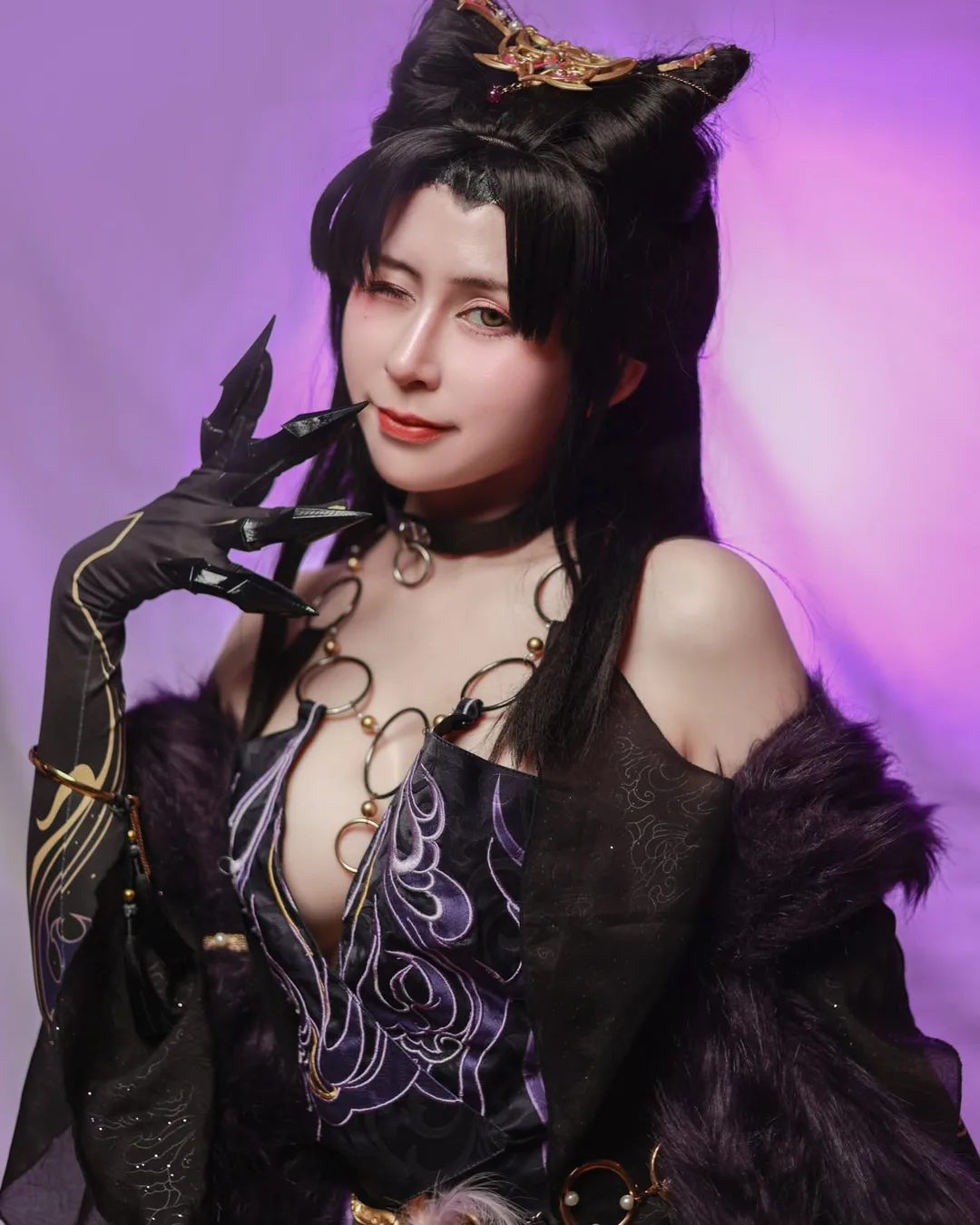 A cosplayer in an ornate black and purple fantasy outfit winks with a clawed hand near her face against a purple backdrop.