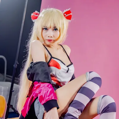 คอสเพลเยอร์ผมบลอนด์แกละสองข้างนั่งบนโต๊ะนักเรียนหน้าฉากสีชมพู สวมถุงเท้ายาวลายทางและเสื้อแจ็กเก็ตสีชมพู