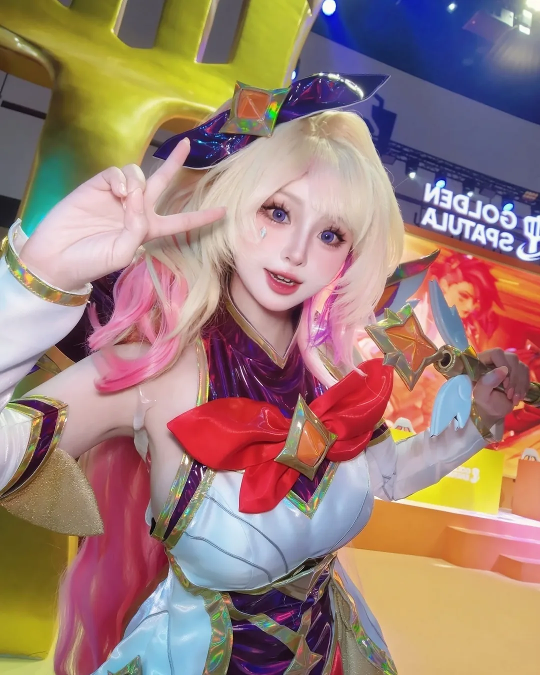 คอสเพลเยอร์ในชุด Star Guardian Seraphine โพสท่าชูสองนิ้วและถือคทาในงานเกม Golden Spatula