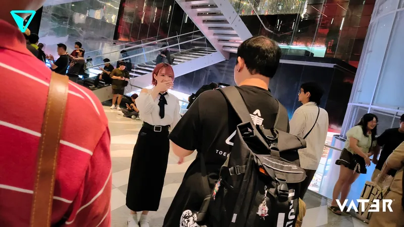 หญิงสาวผมสีชมพูในชุดเสื้อขาวกระโปรงดำยิ้มขณะพูดคุยกับผู้เข้าร่วมงานที่สะพายเป้ในโถงอาคารที่มีผู้คนมากมาย