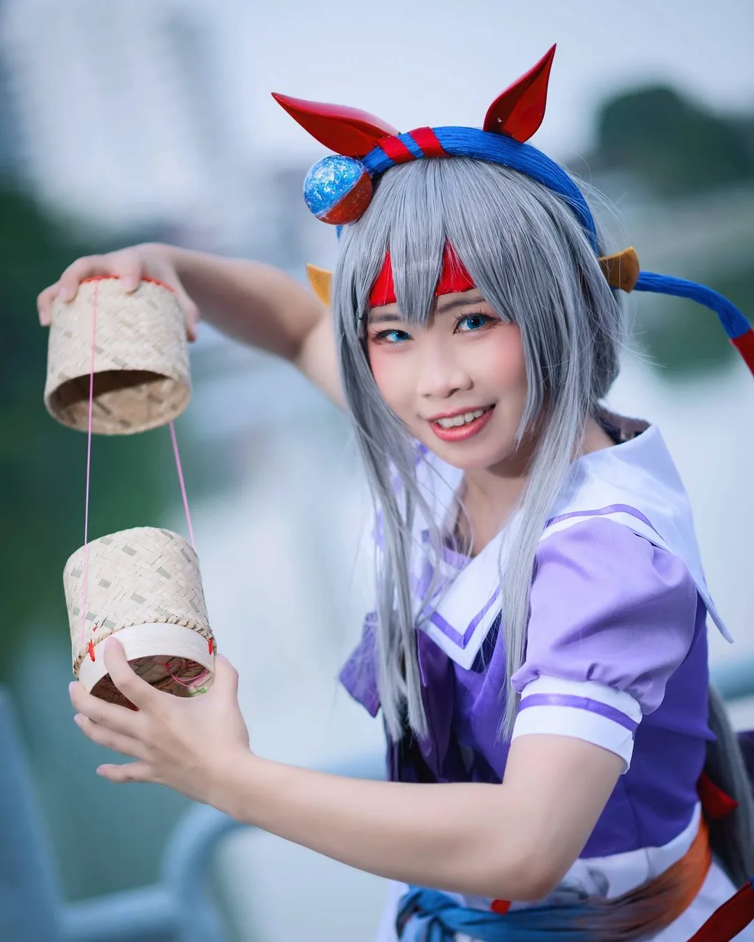 คอสเพลเยอร์ในชุด Tamamo Cross จาก Uma Musume กำลังยิ้มพร้อมถือกระติบข้าวเหนียวสานอยู่กลางแจ้ง