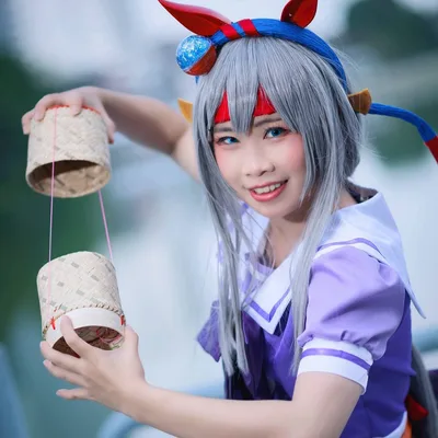 คอสเพลเยอร์ในชุด Tamamo Cross จาก Uma Musume กำลังยิ้มพร้อมถือกระติบข้าวเหนียวสานอยู่กลางแจ้ง