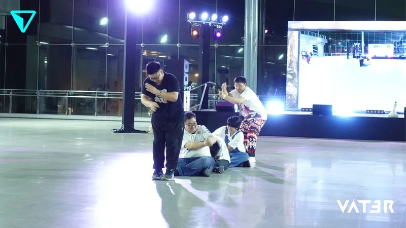 กลุ่มนักแสดงกำลังเต้นและโพสท่าบนพื้นขัดเงาหน้าเวทีที่มีแสงไฟส่องสว่างในงานอีเวนต์ยามค่ำคืน