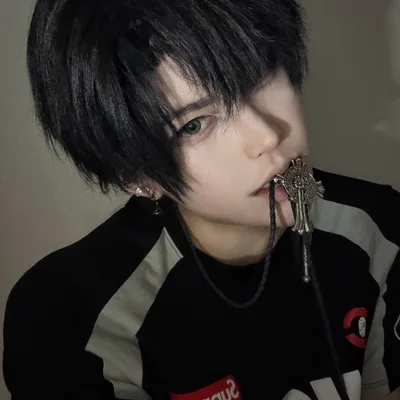 A person with short black hair and light eyes bites the cord of an ornate cross bolo tie while looking at the camera.