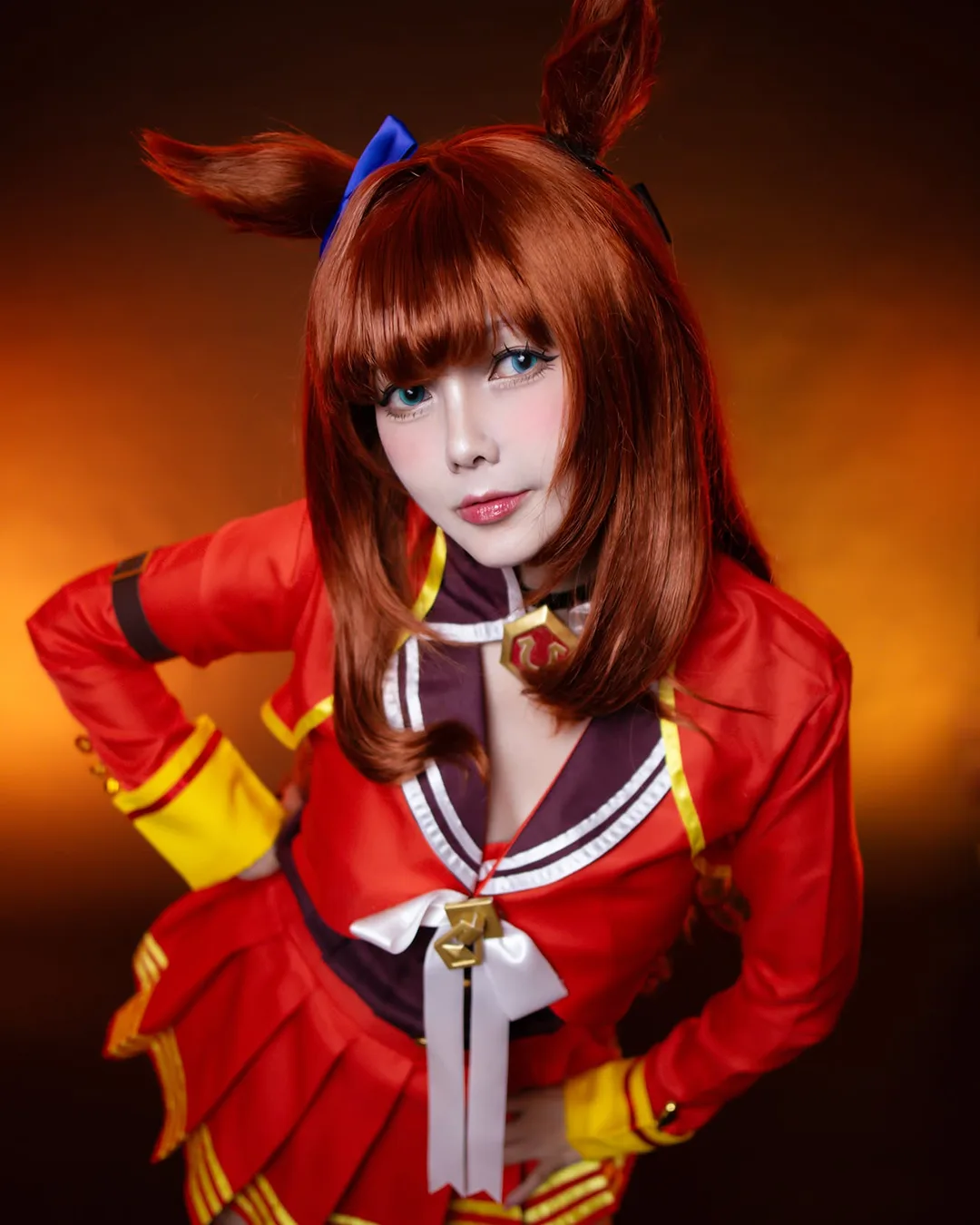 คอสเพลเยอร์แต่งกายเป็น Tokai Teio จาก Uma Musume ในชุดสีแดงทอง โพสท่าโน้มตัวไปข้างหน้าบนพื้นหลังโทนอุ่น