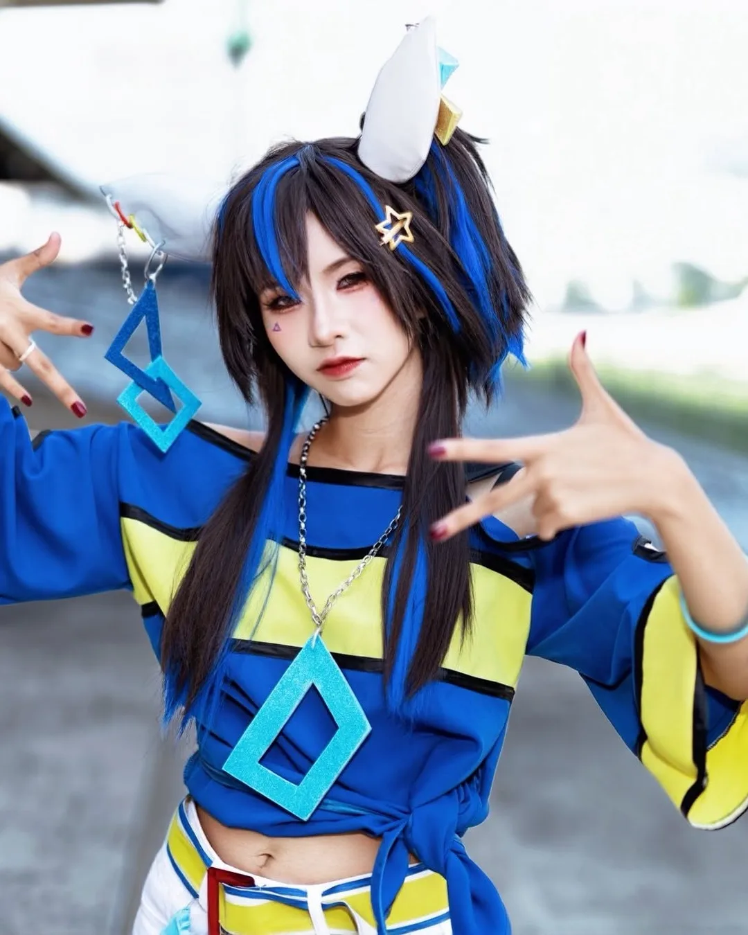 คอสเพลเยอร์แต่งกายเป็น Daitaku Helios จาก Uma Musume Pretty Derby ในเสื้อสีน้ำเงินเหลือง กำลังโพสท่าสไตล์ฮิปฮอปกลางแจ้ง