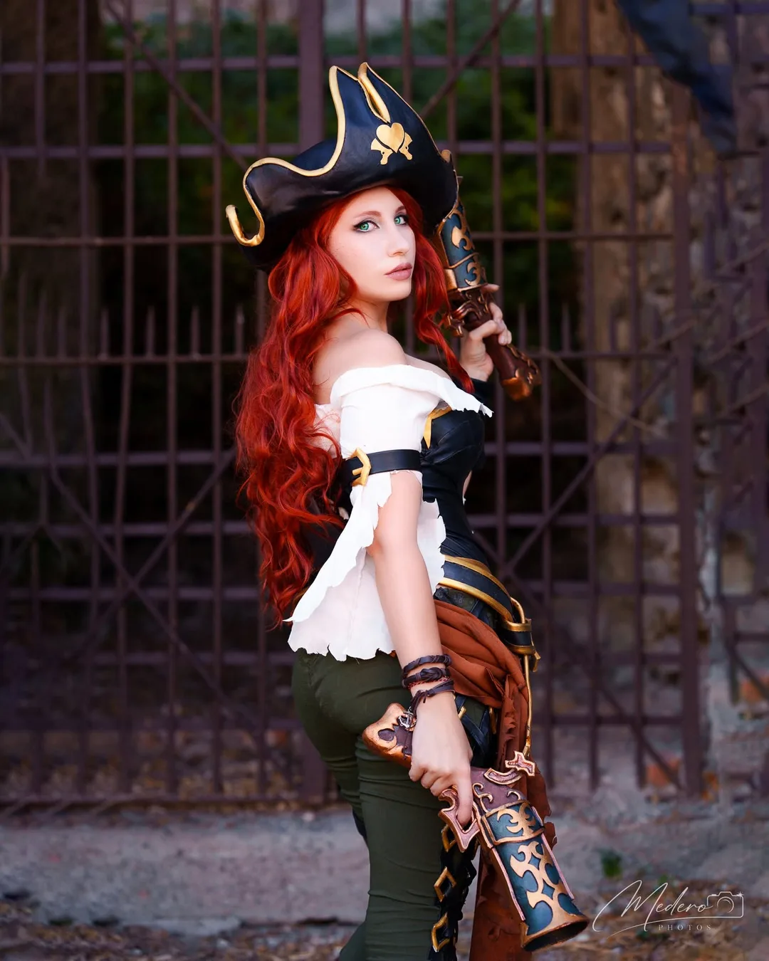 หญิงสาวแต่งคอสเพลย์เป็น Miss Fortune จาก League of Legends ถือปืนพกคู่ลวดลายวิจิตรยืนอยู่หน้าประตูเหล็ก