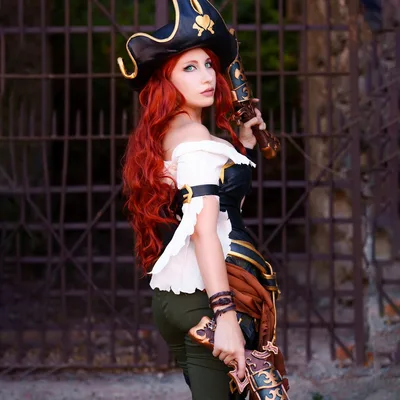 A woman cosplays as Miss Fortune from League of Legends, holding ornate dual pistols in front of an iron gate.