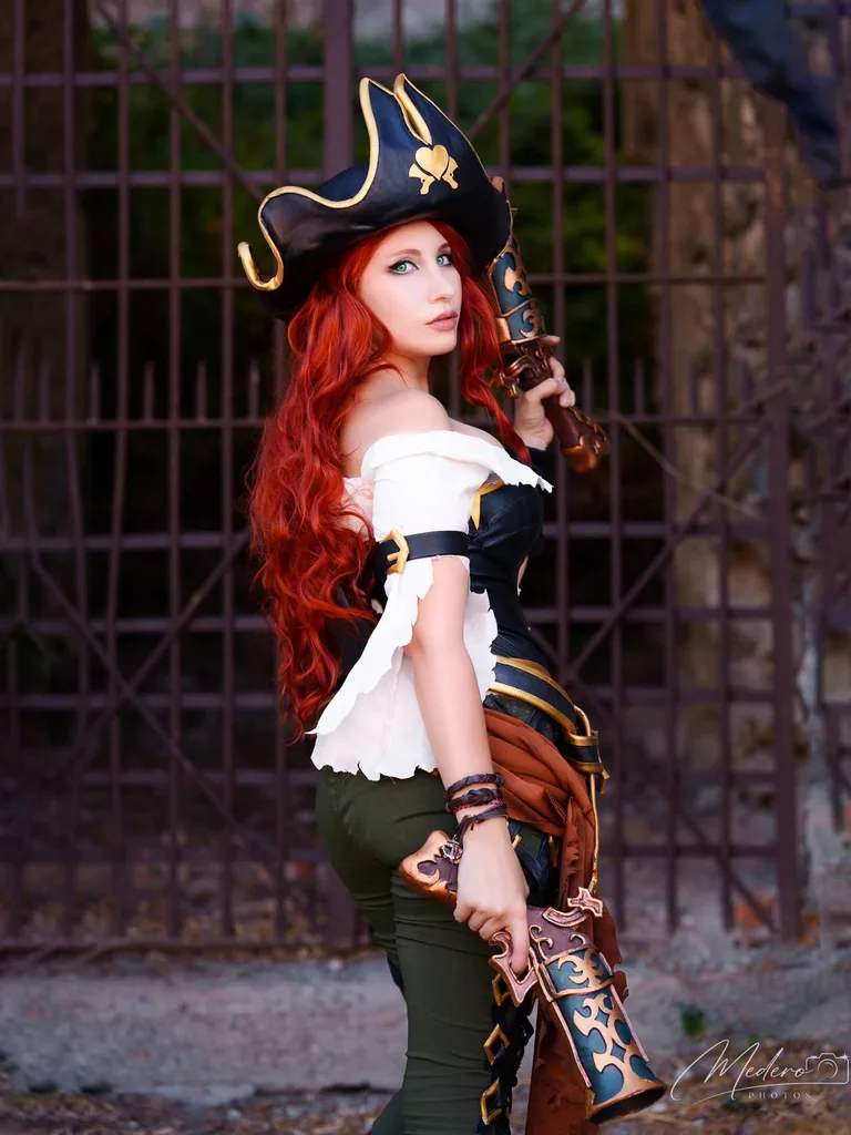 หญิงสาวแต่งคอสเพลย์เป็น Miss Fortune จาก League of Legends ถือปืนพกคู่ลวดลายวิจิตรยืนอยู่หน้าประตูเหล็ก