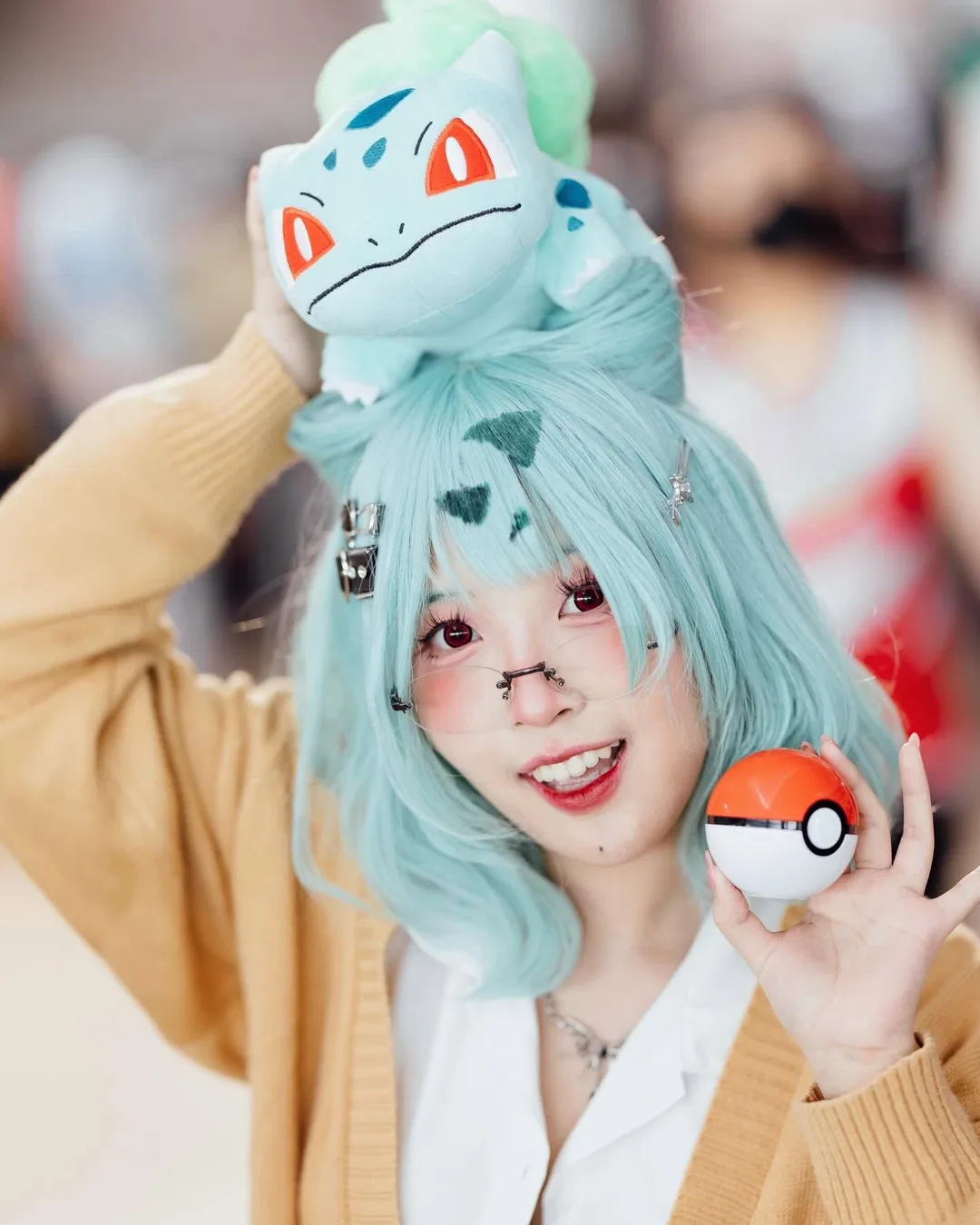 คอสเพลเยอร์ยิ้มแย้มใส่วิกผมสีฟ้าอมเขียว มีตุ๊กตา Bulbasaur วางอยู่บนศีรษะ และถือ Poké Ball ไว้ในมือ