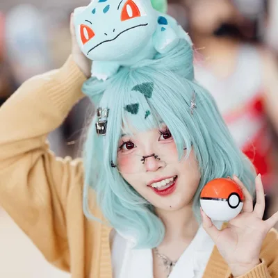 คอสเพลเยอร์ยิ้มแย้มใส่วิกผมสีฟ้าอมเขียว มีตุ๊กตา Bulbasaur วางอยู่บนศีรษะ และถือ Poké Ball ไว้ในมือ