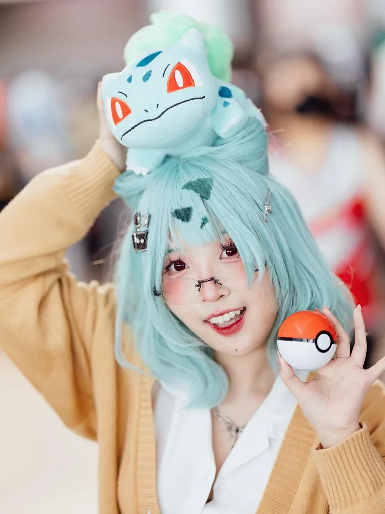 คอสเพลเยอร์ยิ้มแย้มใส่วิกผมสีฟ้าอมเขียว มีตุ๊กตา Bulbasaur วางอยู่บนศีรษะ และถือ Poké Ball ไว้ในมือ
