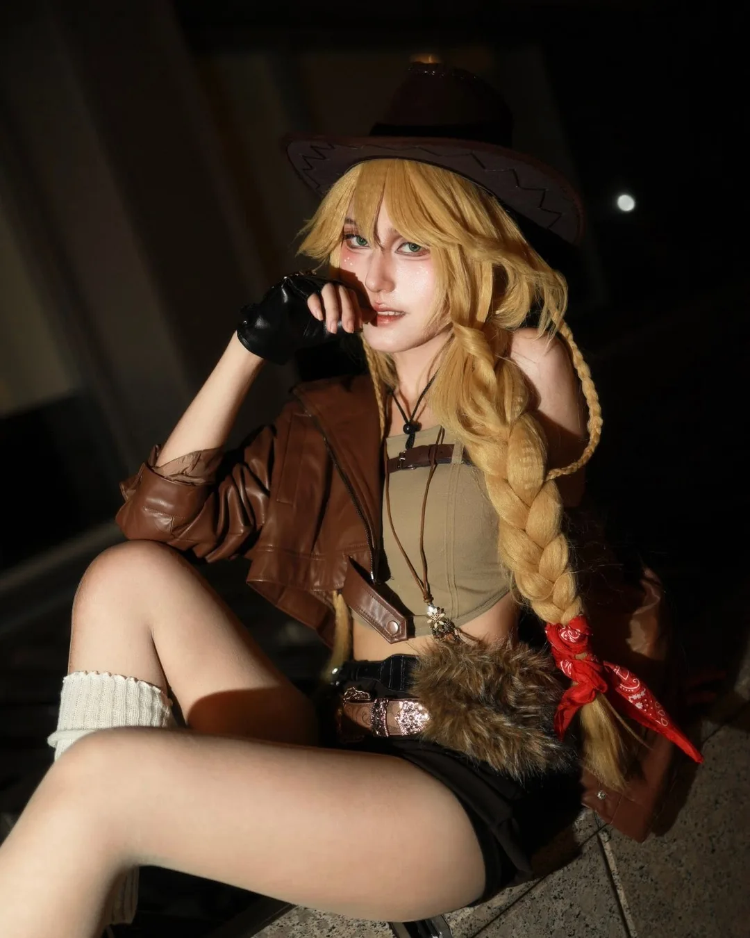 A cosplayer with a blonde braid and brown cowboy hat poses outdoors at night wearing a cropped leather jacket and shorts.