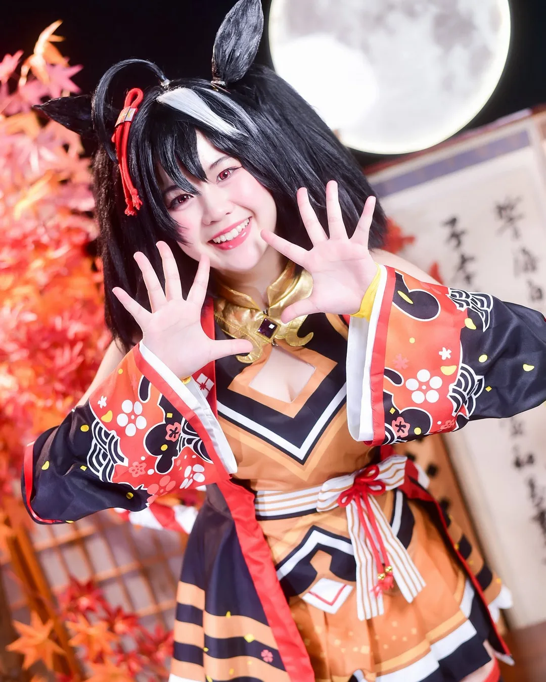 คอสเพลเยอร์ในชุด Kitasan Black จาก Uma Musume ยิ้มสดใสและยกสองมือขึ้นโพสท่าหน้าฉากหลังใบไม้เปลี่ยนสีและพระจันทร์เต็มดวง