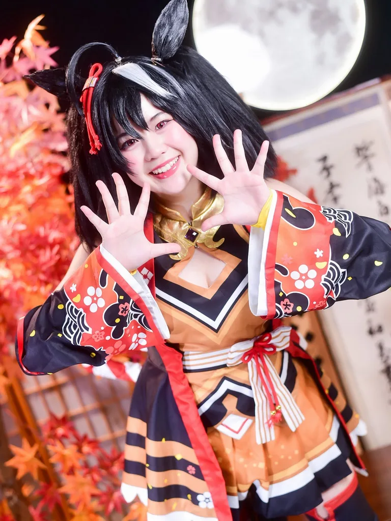 คอสเพลเยอร์ในชุด Kitasan Black จาก Uma Musume ยิ้มสดใสและยกสองมือขึ้นโพสท่าหน้าฉากหลังใบไม้เปลี่ยนสีและพระจันทร์เต็มดวง