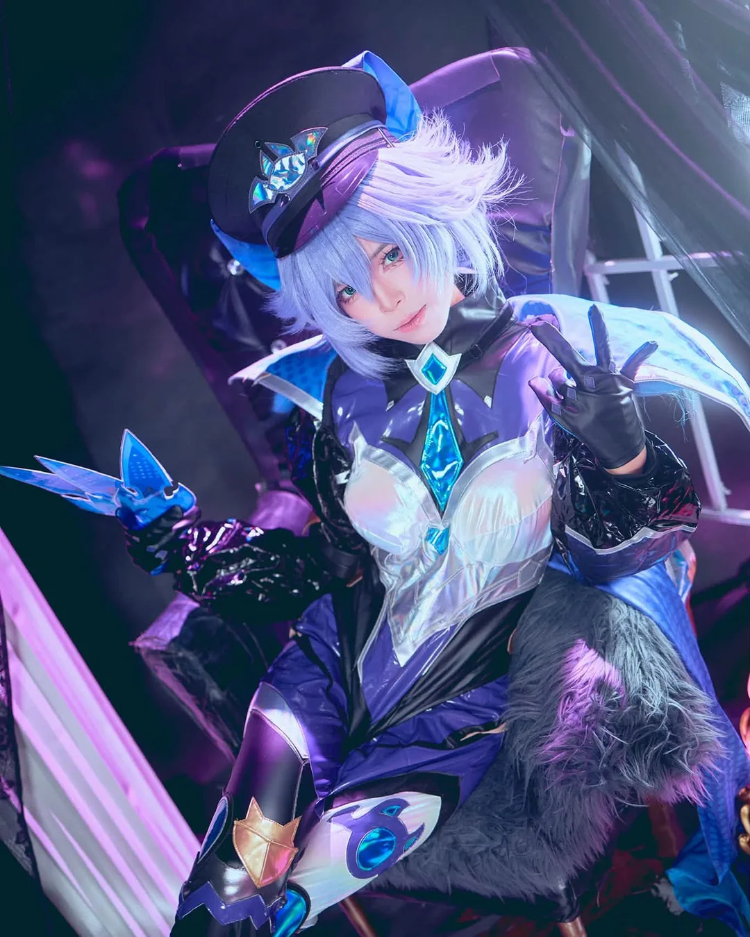A cosplayer with short lavender hair in a blue and black fantasy officer uniform sits on a chair, making a peace sign.