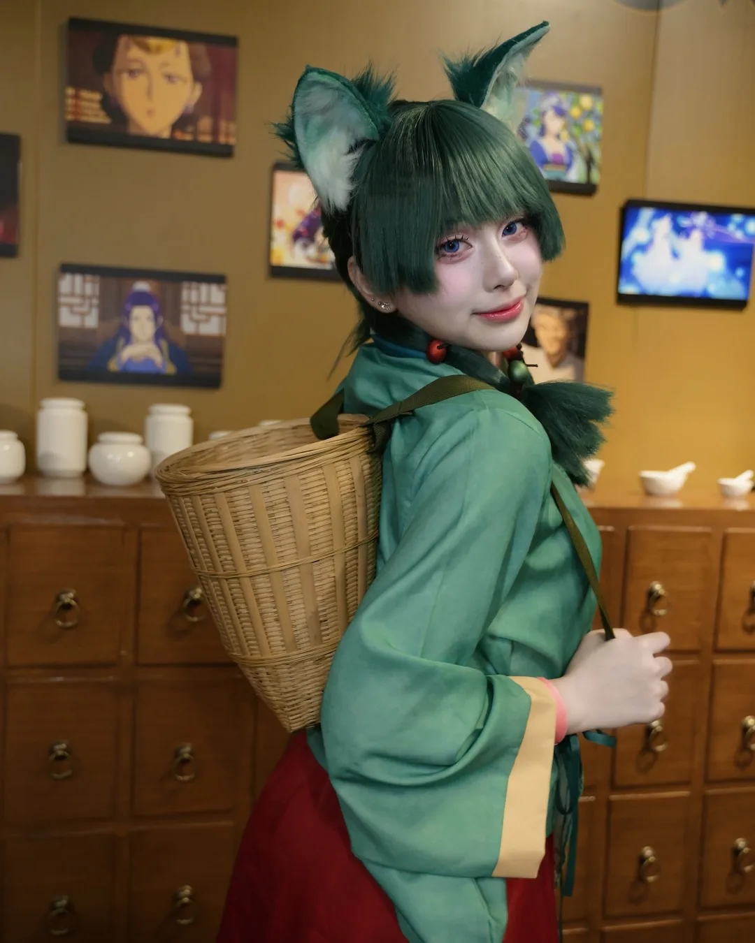 คอสเพลเยอร์แต่งกายเป็น Maomao จาก The Apothecary Diaries สวมหูแมวและสะพายตะกร้าสาน ยืนอยู่หน้าตู้ลิ้นชักยาจีน