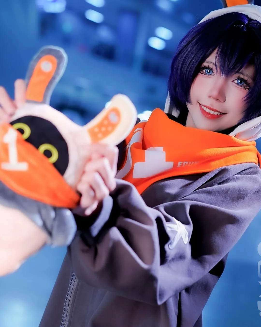 คอสเพลเยอร์แต่งเป็น Belle จาก Zenless Zone Zero ยิ้มและถือตุ๊กตา Bangboo Eous ในงานอีเวนต์ในร่ม