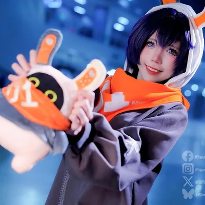 A cosplayer dressed as Belle from Zenless Zone Zero smiles while holding an Eous Bangboo plush at an indoor event.