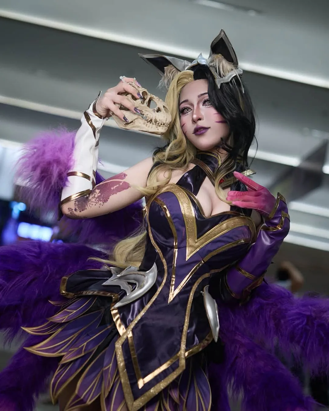 คอสเพลเยอร์ในชุด Coven Ahri โพสท่าถือหน้ากากกะโหลกสัตว์พร้อมหางฟูสีม่วงในงานจัดแสดงในร่ม