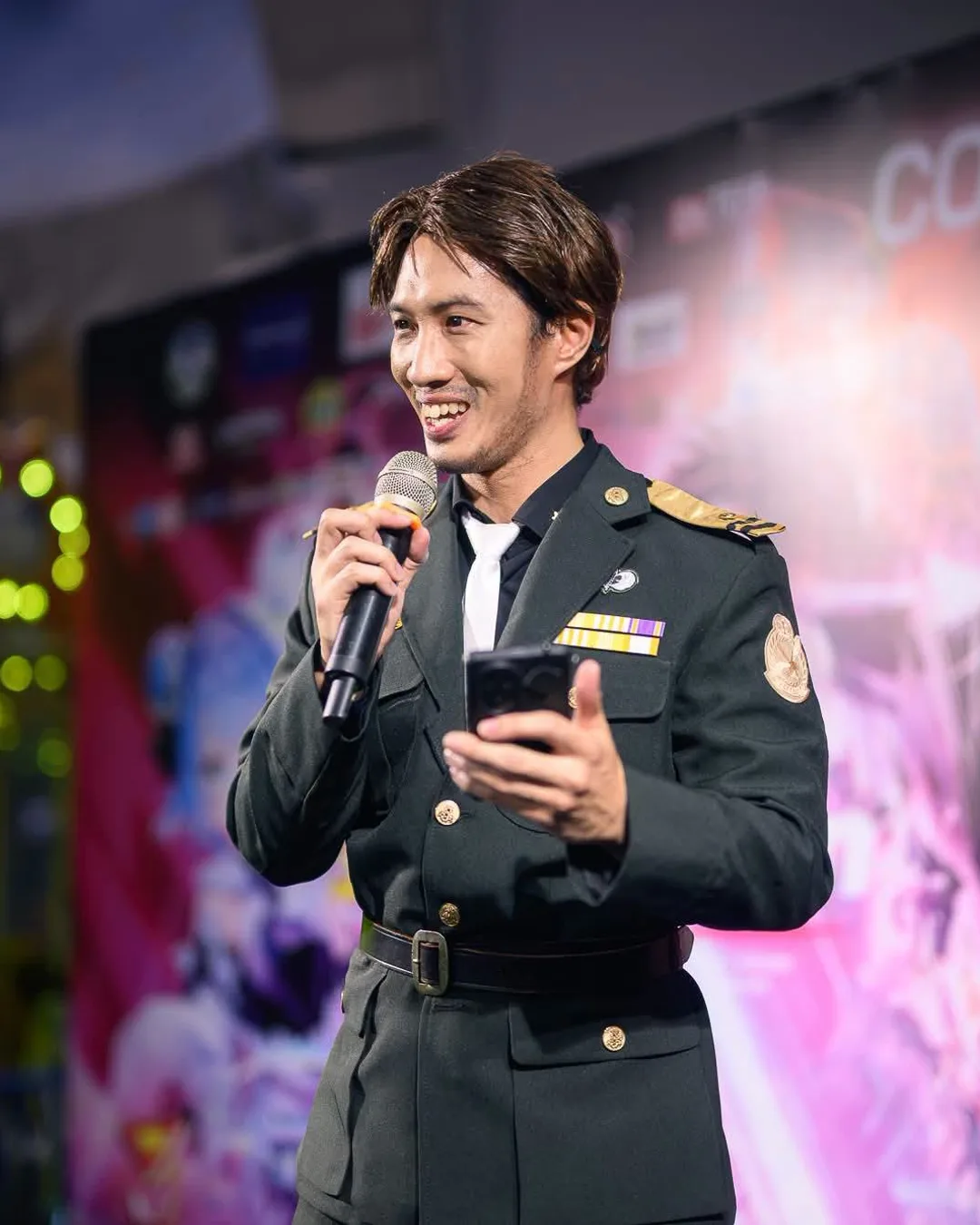 ชายในชุดคอสเพลย์เครื่องแบบทหารพูดใส่ไมโครโฟนพร้อมถือสมาร์ตโฟนบนเวทีงานอีเวนต์คอสเพลย์