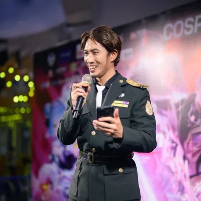 ชายในชุดคอสเพลย์เครื่องแบบทหารพูดใส่ไมโครโฟนพร้อมถือสมาร์ตโฟนบนเวทีงานอีเวนต์คอสเพลย์