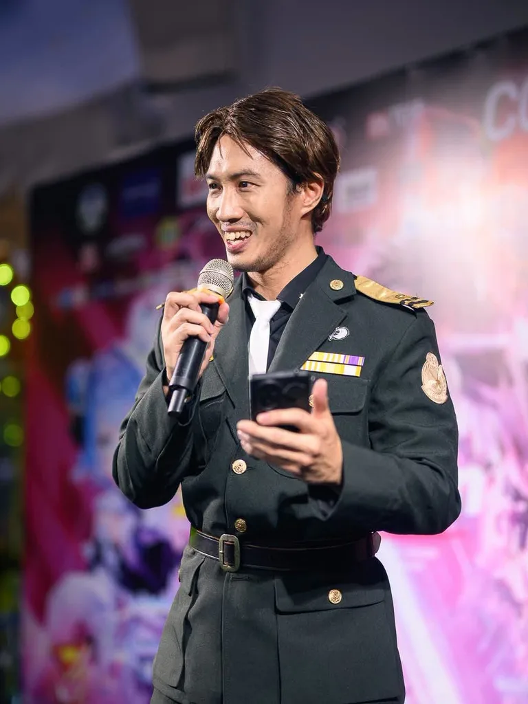 ชายในชุดคอสเพลย์เครื่องแบบทหารพูดใส่ไมโครโฟนพร้อมถือสมาร์ตโฟนบนเวทีงานอีเวนต์คอสเพลย์