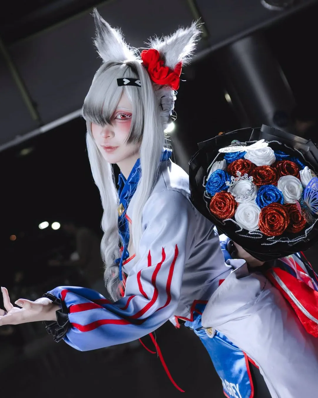 คอสเพลเยอร์ผมสีเงินติดหูสัตว์ยื่นมือออกมาข้างหนึ่ง พร้อมถือช่อดอกกุหลาบสีแดง น้ำเงิน และขาวในงานอีเวนต์