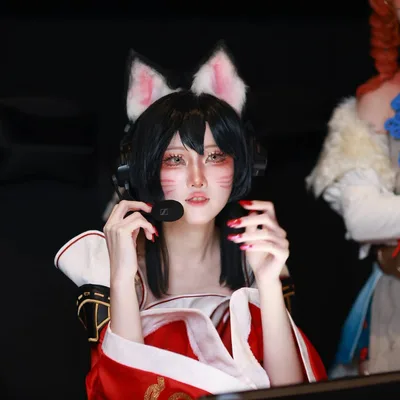 คอสเพลเยอร์ในชุด Ahri สวมหูจิ้งจอกและชุดหูฟัง กำลังปรับไมโครโฟนขณะนั่งอยู่ที่โต๊ะเล่นเกม