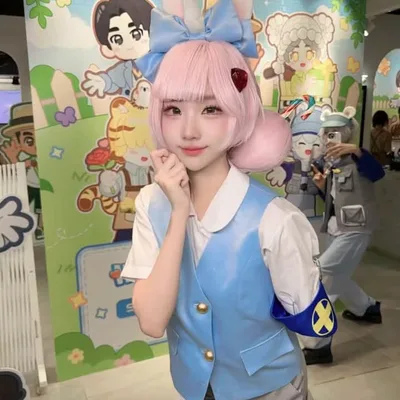 คอสเพลเยอร์ผมสีชมพูสวมหูกระต่ายโพสท่าในเสื้อกั๊กสีฟ้าอ่อนและกางเกงขาสั้น หน้าฉากหลังภาพวาดการ์ตูนในงานอีเวนต์