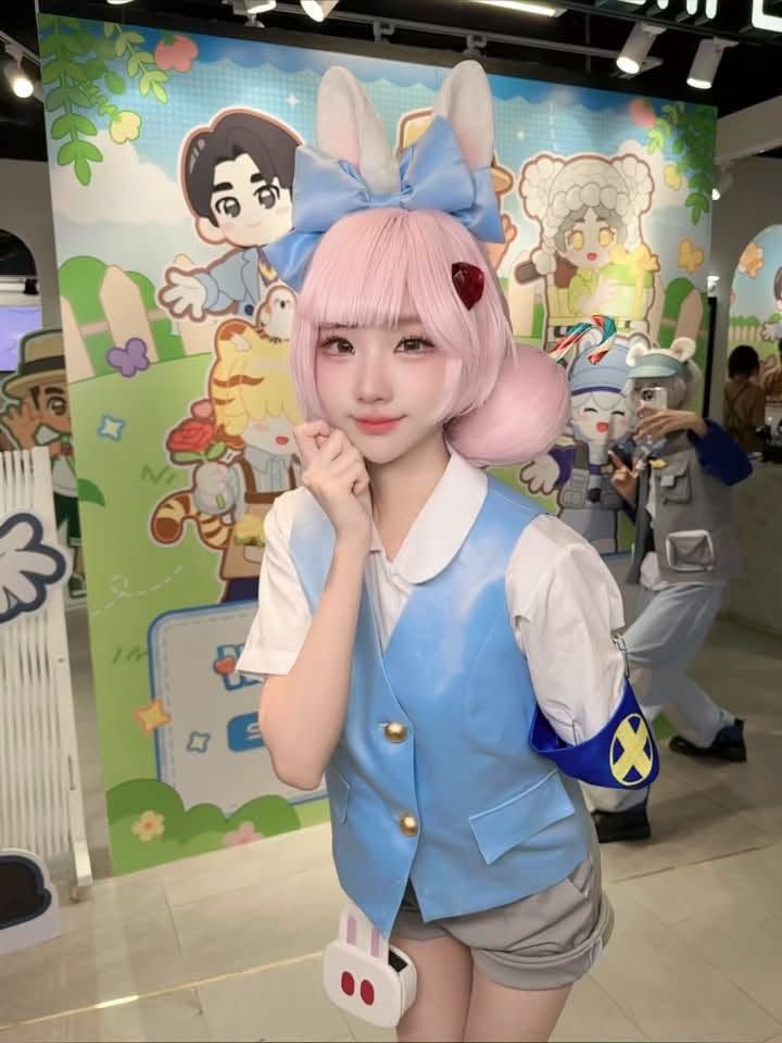 คอสเพลเยอร์ผมสีชมพูสวมหูกระต่ายโพสท่าในเสื้อกั๊กสีฟ้าอ่อนและกางเกงขาสั้น หน้าฉากหลังภาพวาดการ์ตูนในงานอีเวนต์