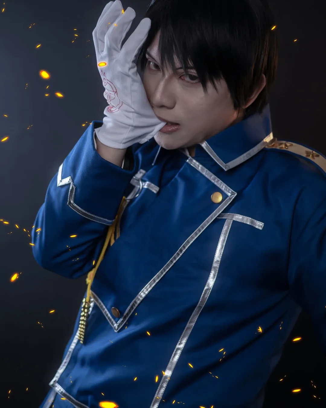 คอสเพลเยอร์แต่งกายเป็น Roy Mustang จาก Fullmetal Alchemist สวมเครื่องแบบทหารสีน้ำเงินและกัดถุงมือสีขาวท่ามกลางประกายไฟ
