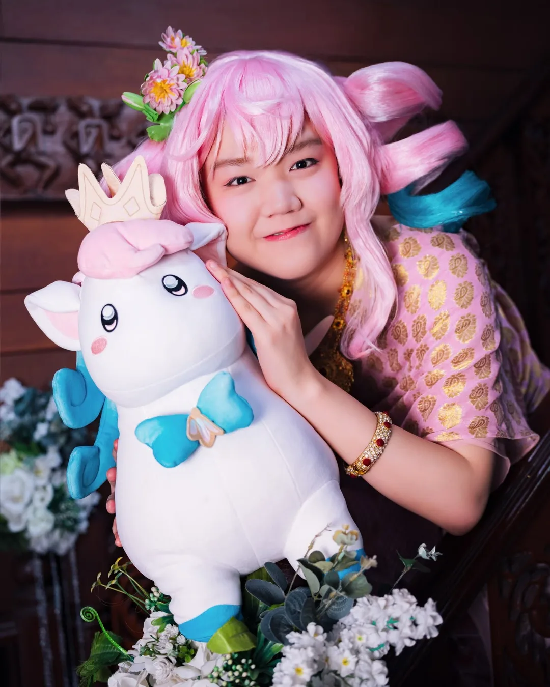 คอสเพลเยอร์ผมสีชมพูยิ้มหวานขณะกอดตุ๊กตาการ์ตูนสีขาวตัวใหญ่ข้างราวบันไดไม้ที่ประดับด้วยดอกไม้