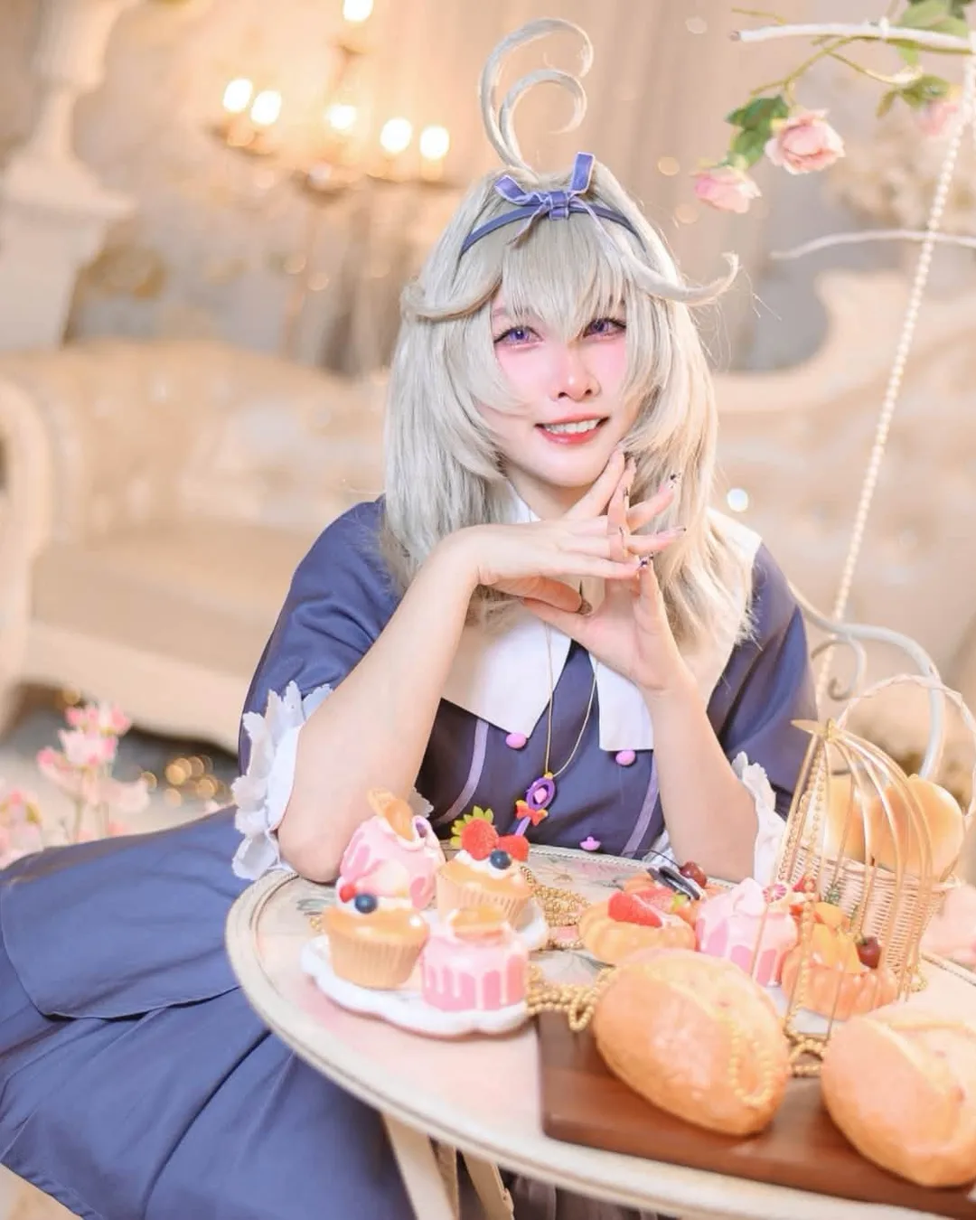 A cosplayer dressed as Robin from Honkai: Star Rail sits smiling at a tea table covered in pastries and flowers in an elegant white room.